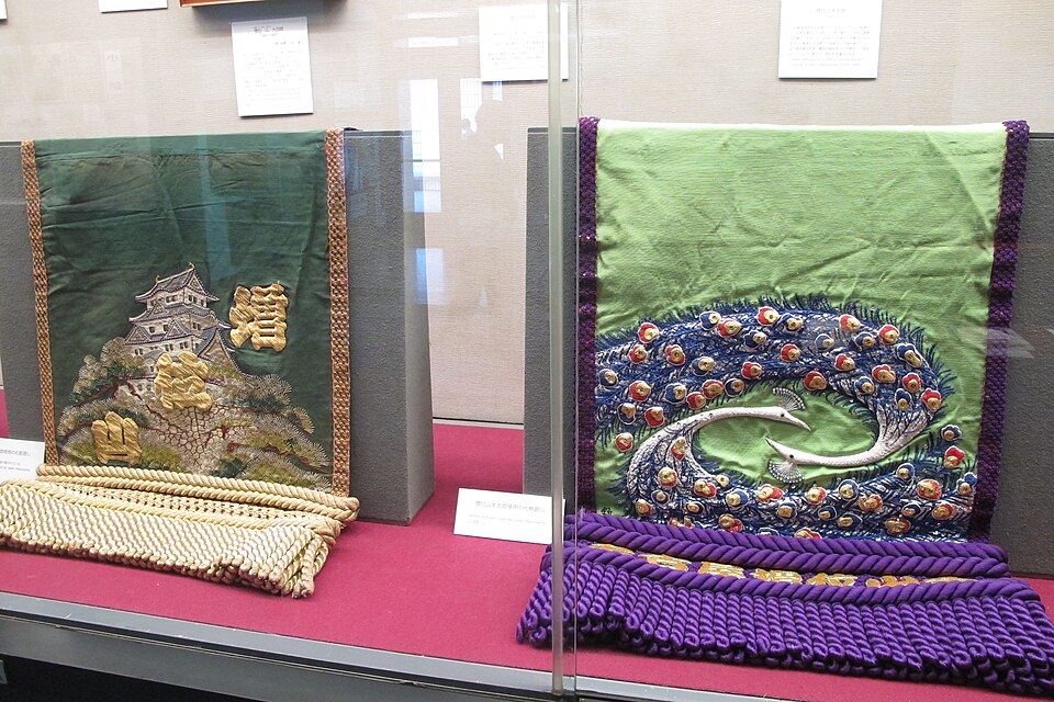 Two embroidered ceremonial sumo aprons displayed in a museum case, one green with a castle, one with a peacock.