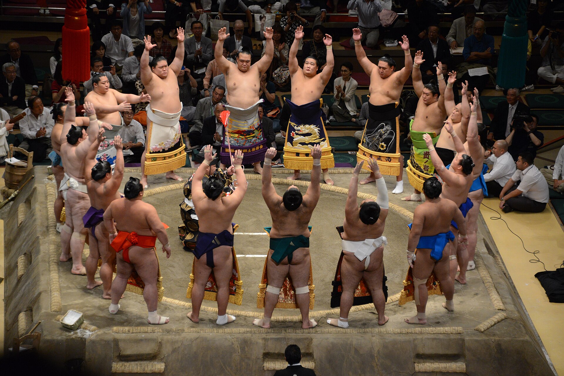 A full ring of wrestlers in embroidered aprons with arms raised.