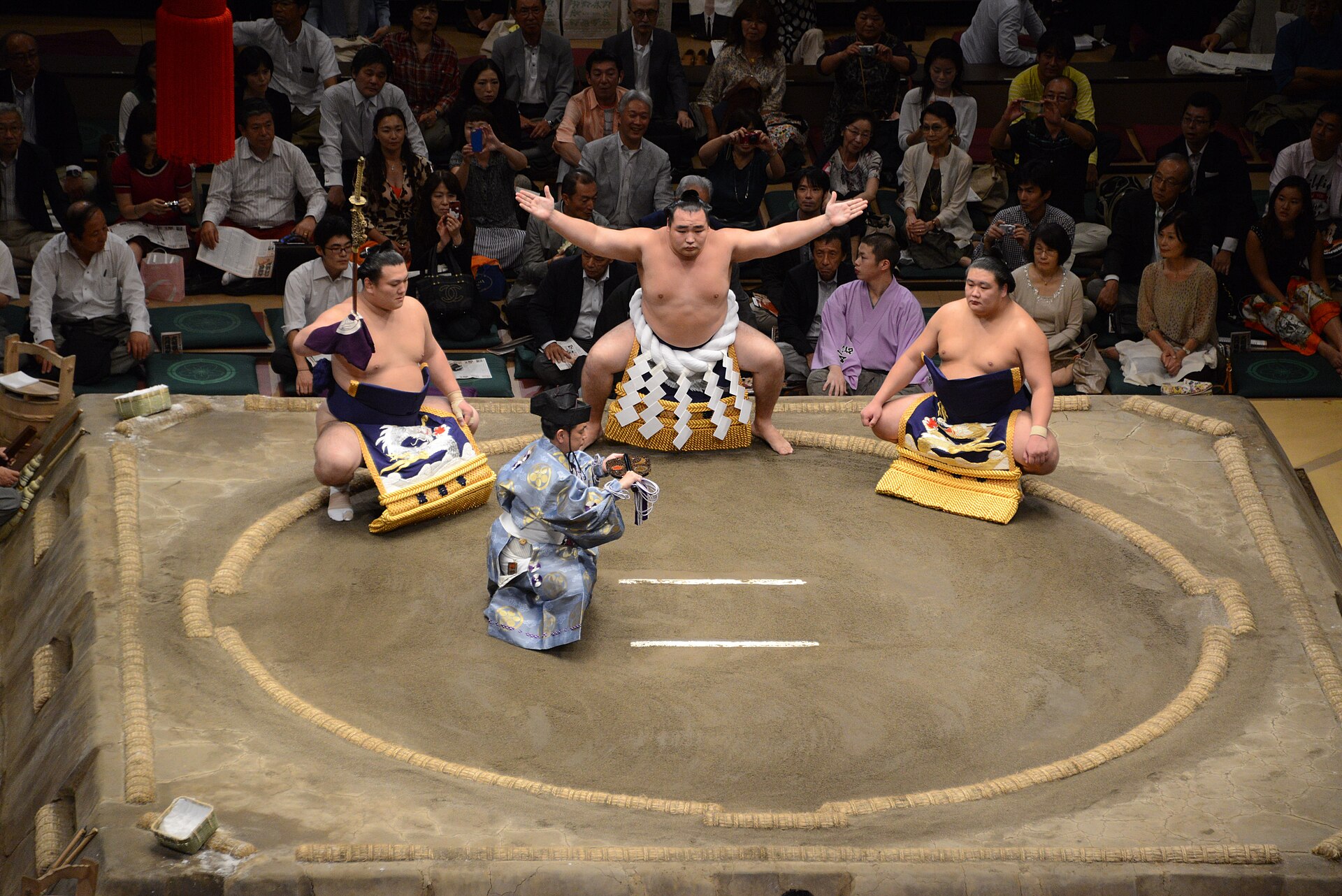 A yokozuna in the white tsuna rope, arms spread, flanked by two attendants in the ring.